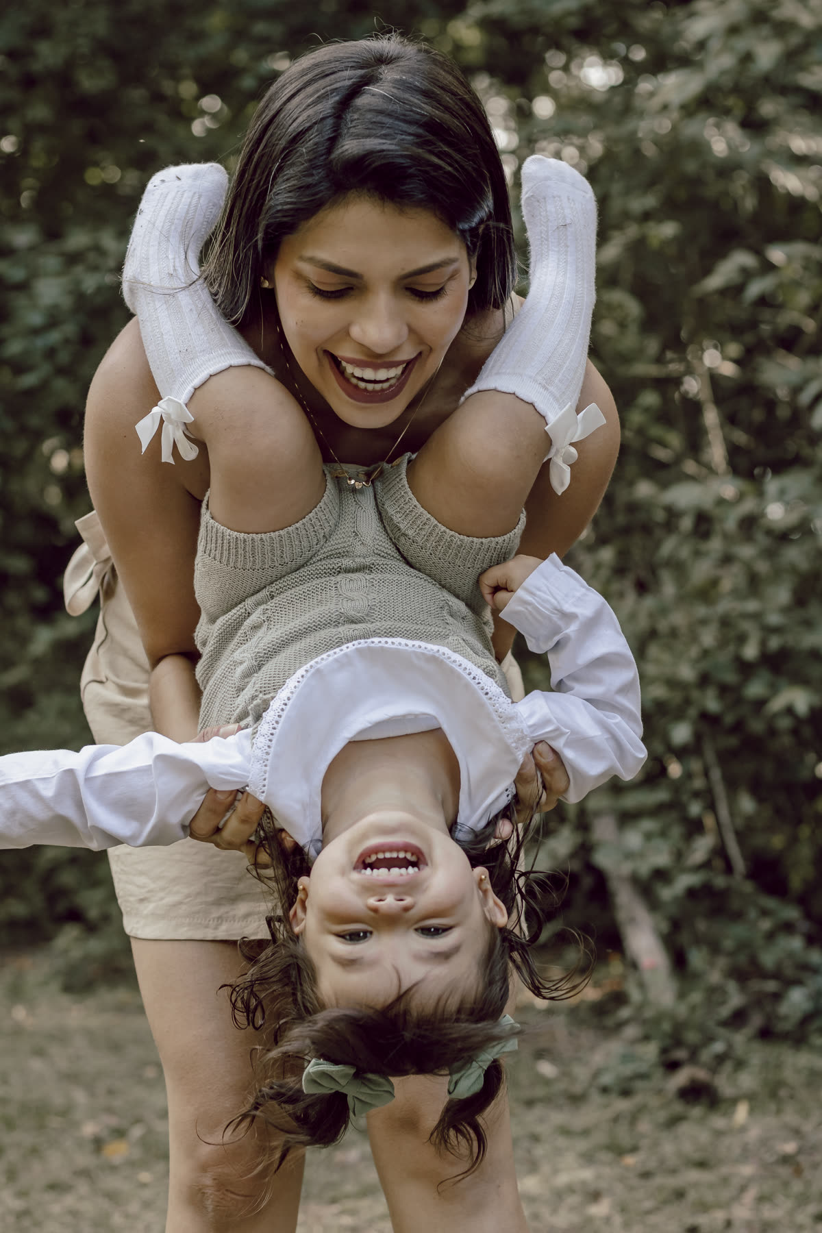 Mulher sorrindo segura a menina de cabeça para baixo num jardim, as duas rindo.