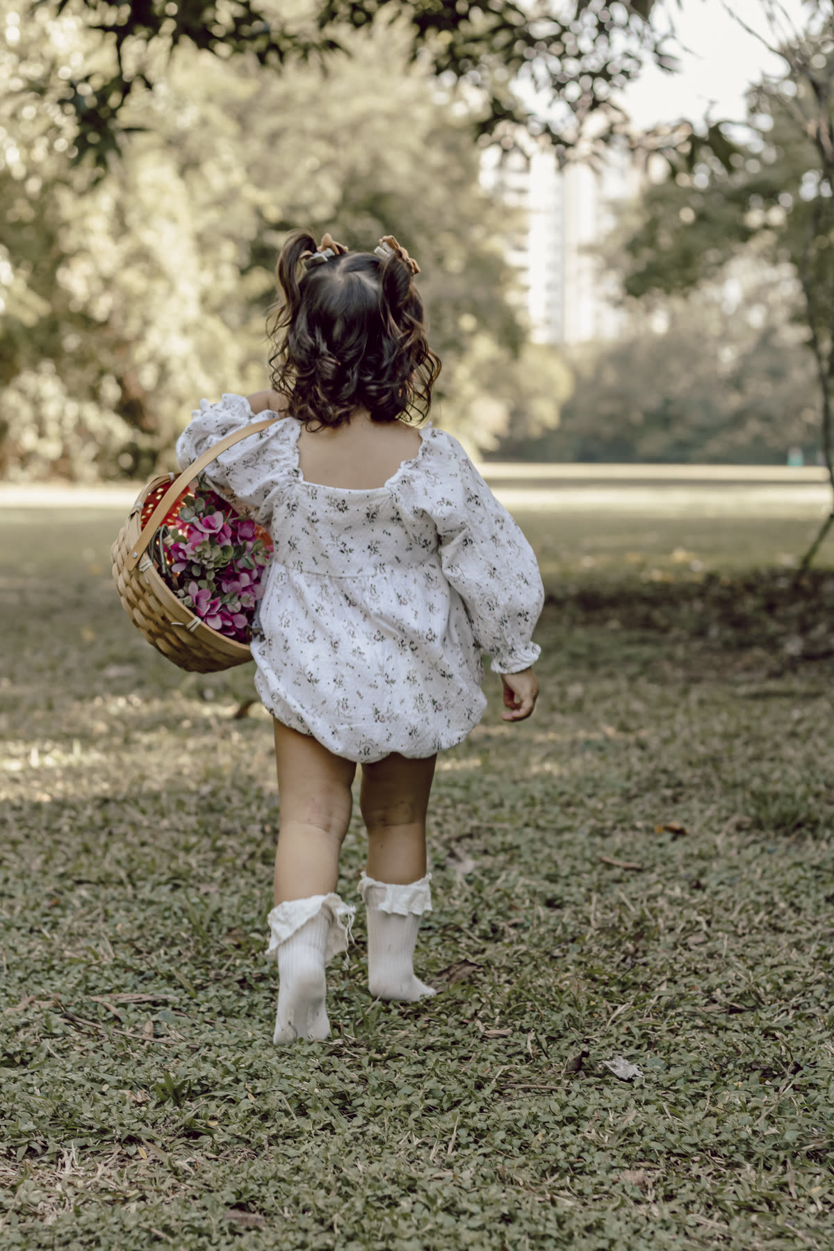 Menina pequena de vestido florido caminha pela grama carregando uma cestinha de flores.