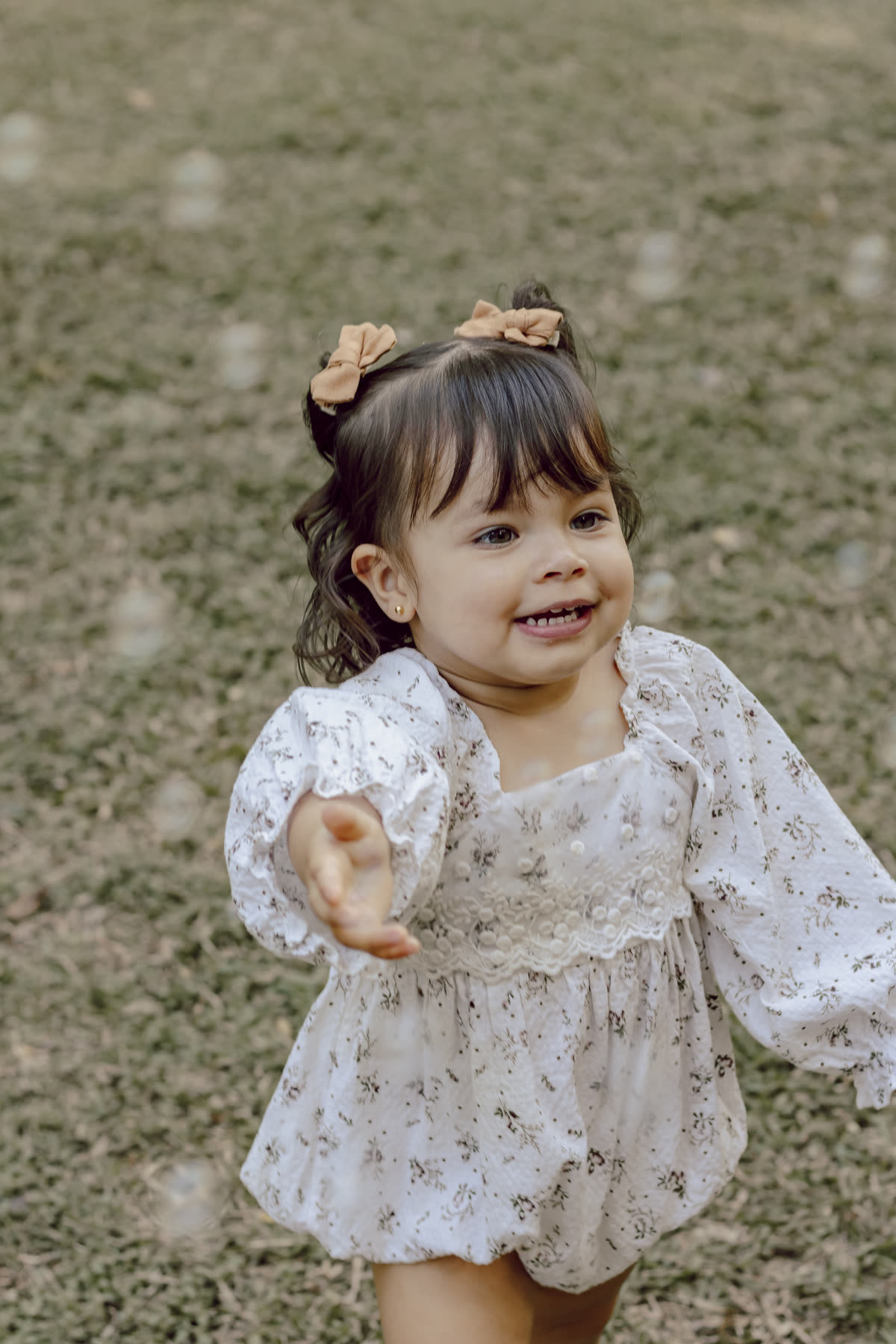 Menina pequena de vestido florido corre pela grama, sorrindo, com a mão estendida.