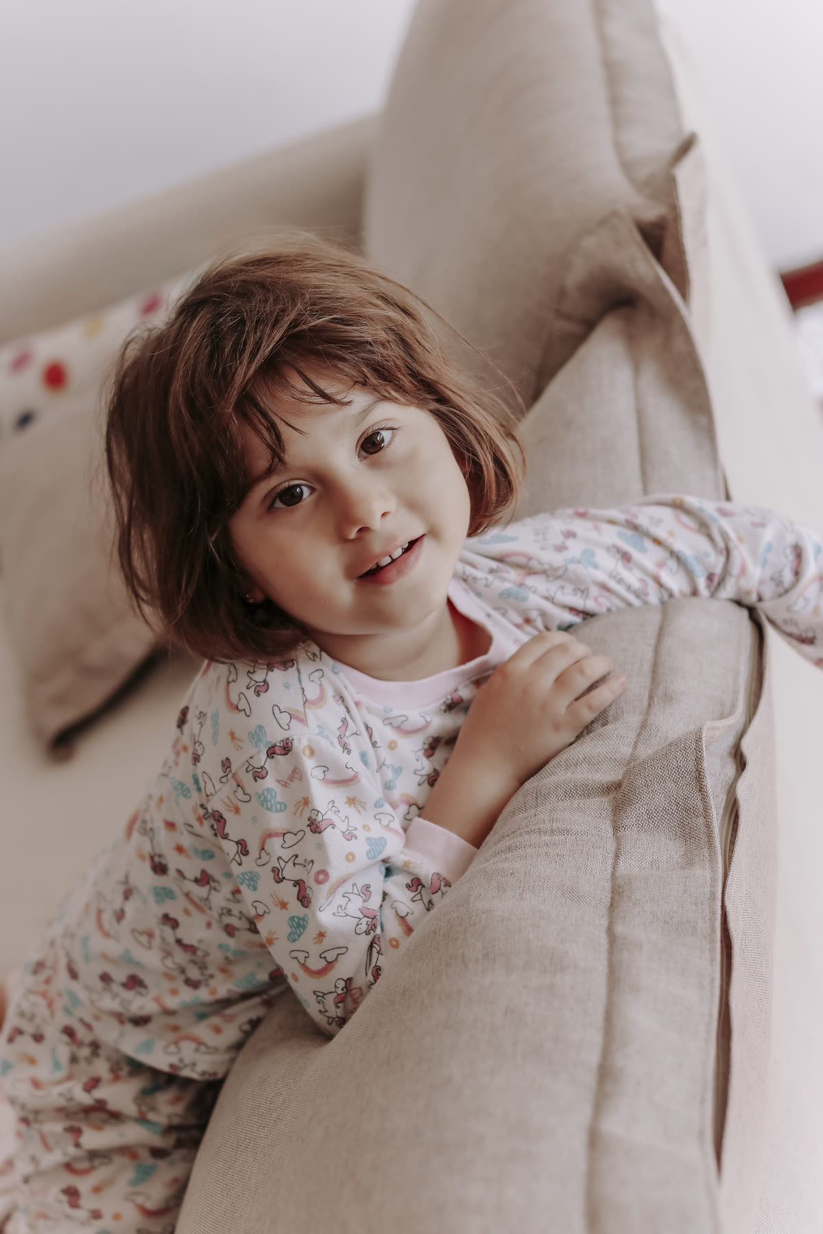 Menina de pijama florido debruçada no braço do sofá, olhando para a câmera.