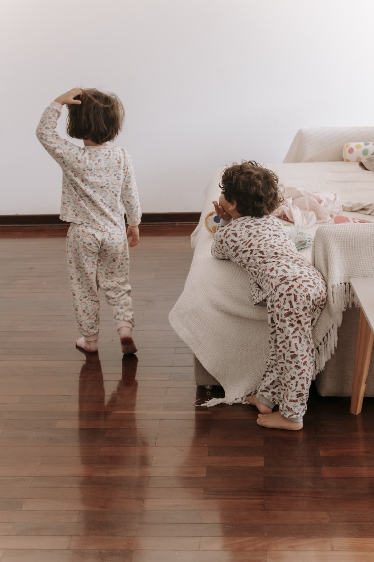 Duas crianças de pijama brincando na sala de piso de madeira, uma delas debruçada no sofá.