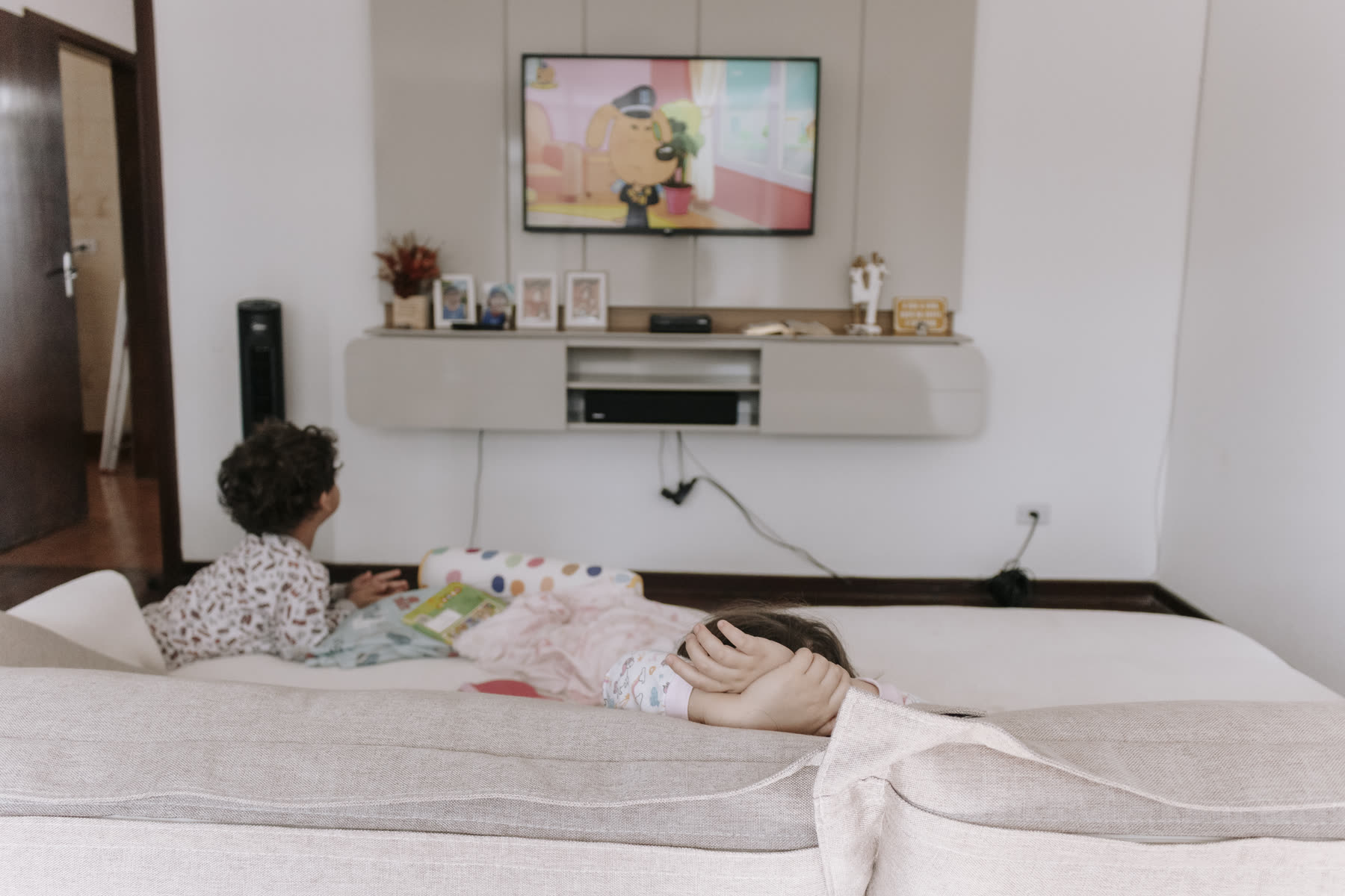 Duas crianças de pijama deitadas na cama de casal, assistindo desenho na televisão do quarto.