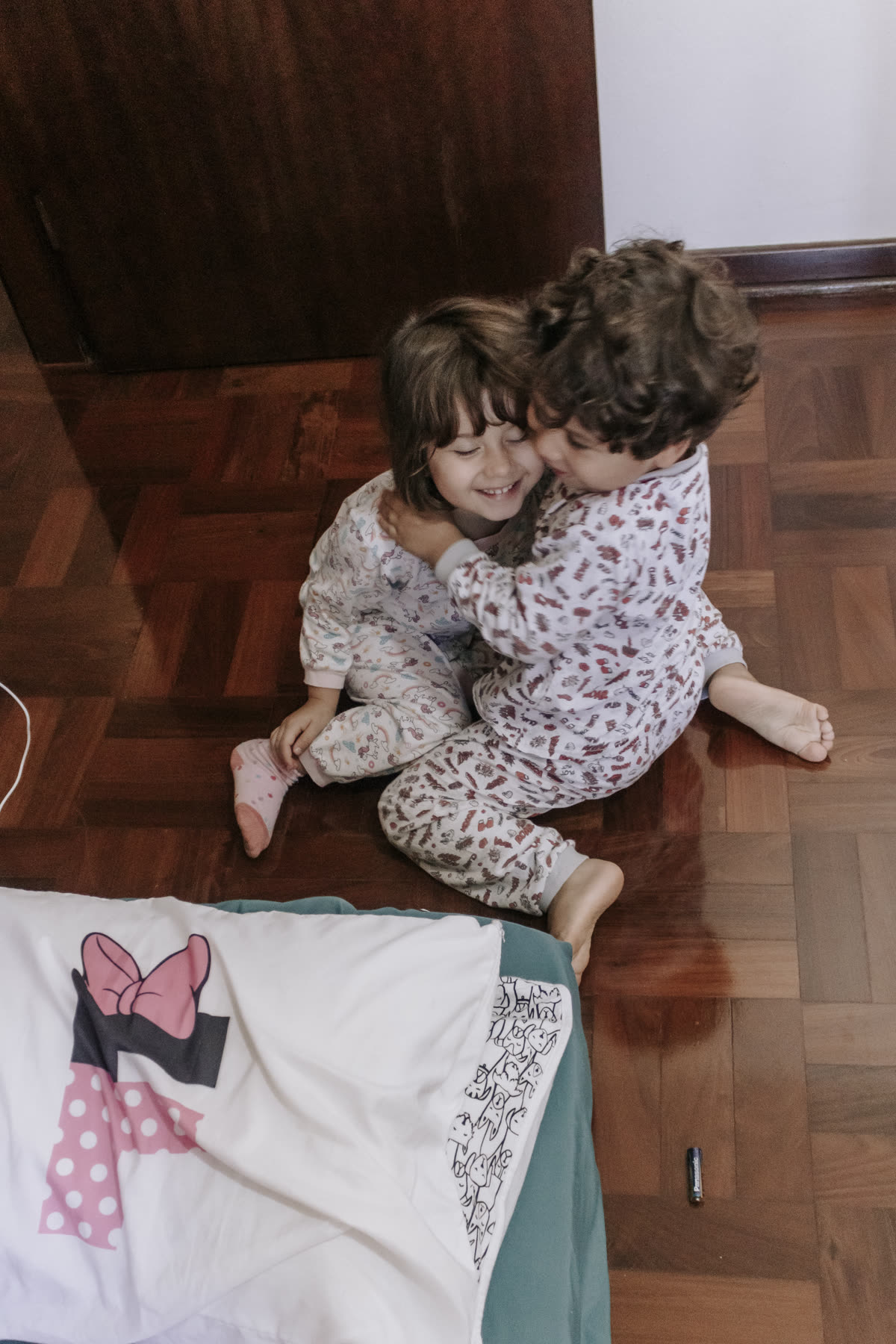 Dois irmãos de pijama abraçados no chão de taco da sala.
