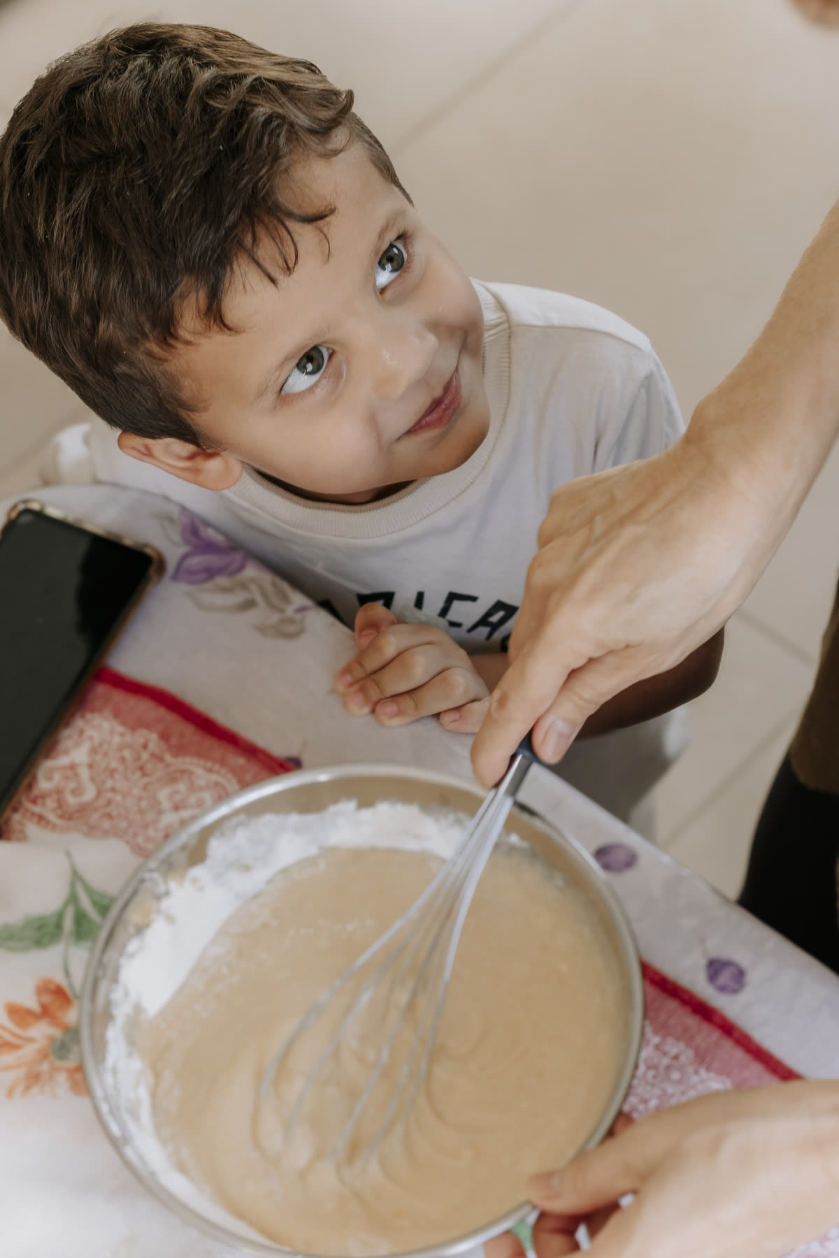 Menino olha para cima enquanto um adulto bate a massa de bolo numa tigela.