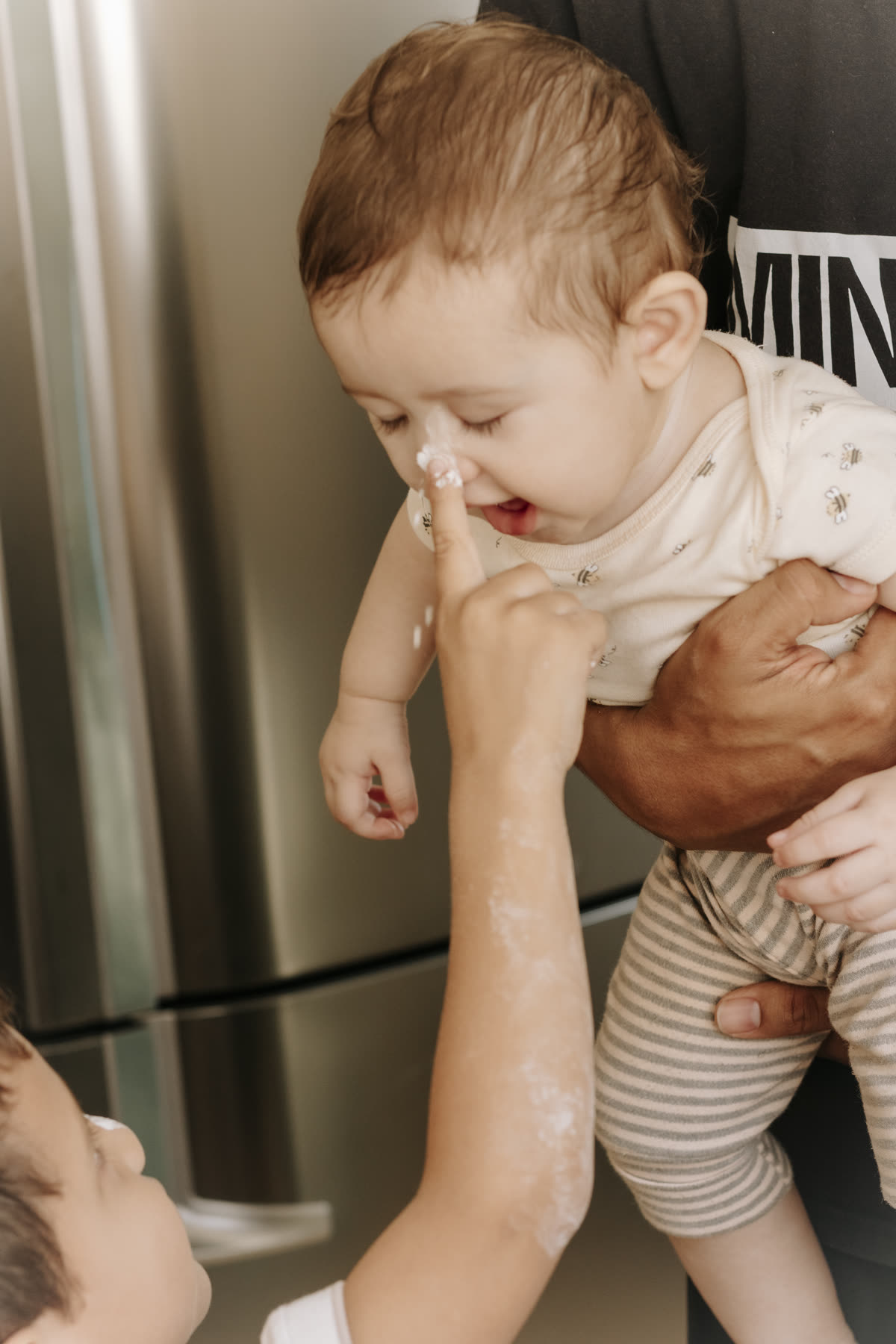Bebê no colo, com farinha no nariz, enquanto alguém encosta o dedo no rosto dele.