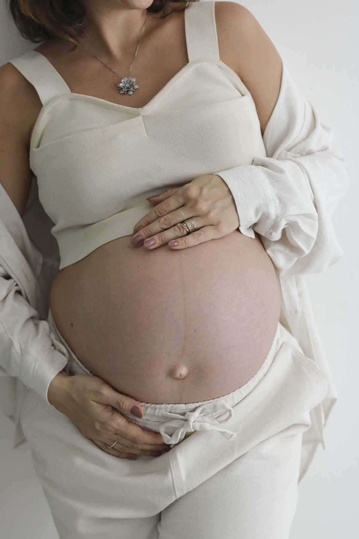 Mãos da gestante apoiadas na barriga, sobre roupa clara.
