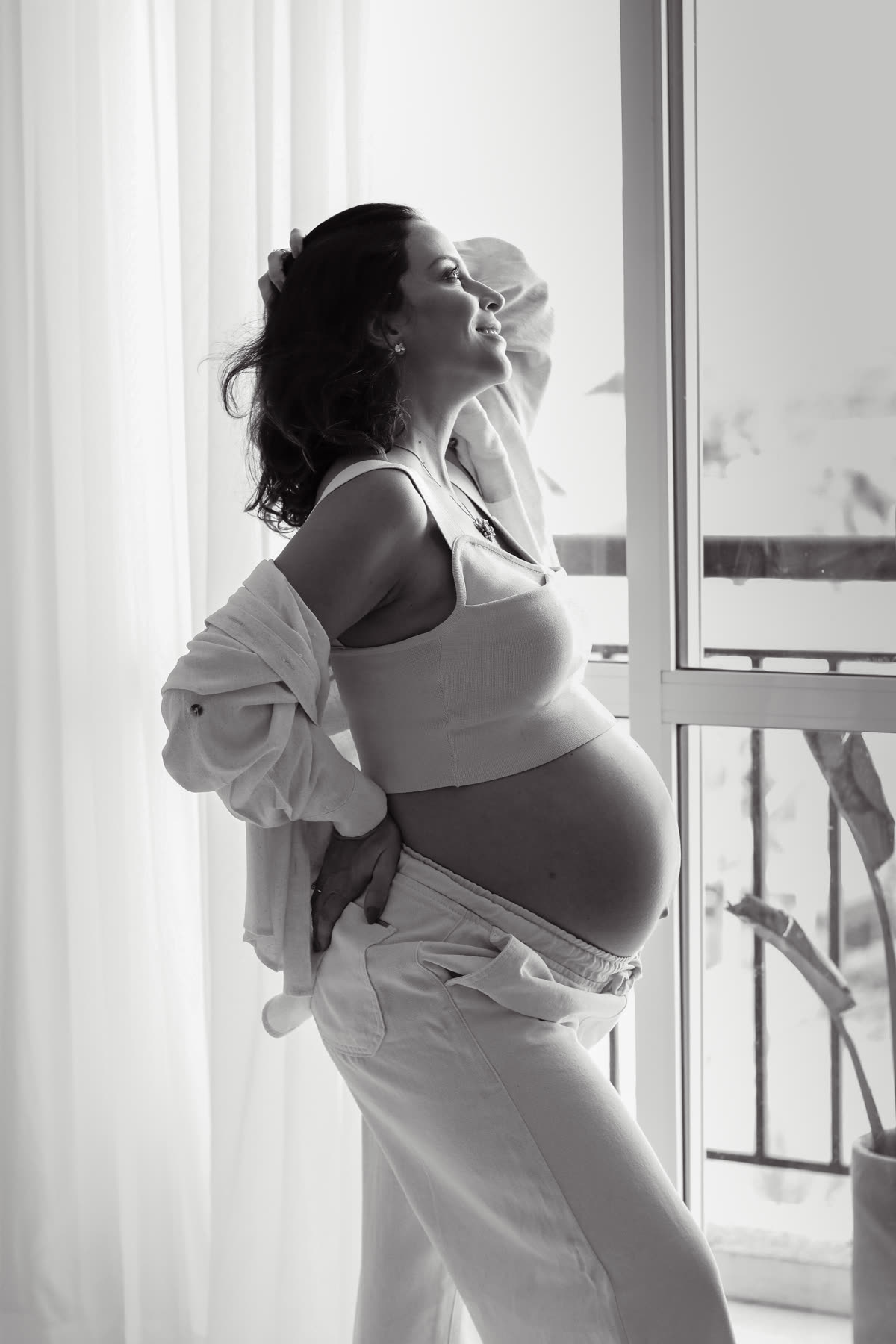 Gestante em preto e branco, de perfil junto à janela, com a mão na barriga.